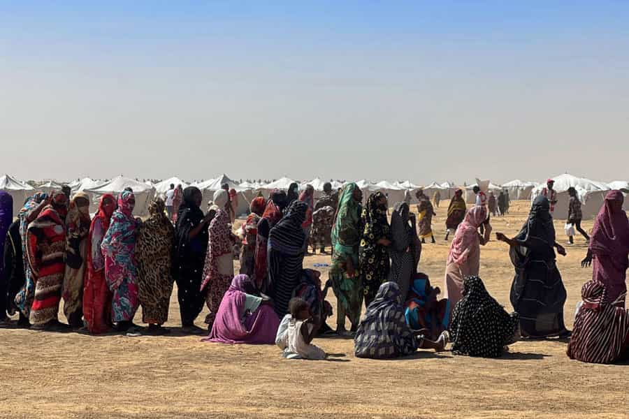 در میان انبوهی از چادرهای بنا شده در صحرا، ده‌ها زن سودانی در صف ایستاده‌اند تا کمک‌های توزیع‌شده را دریافت کنند.