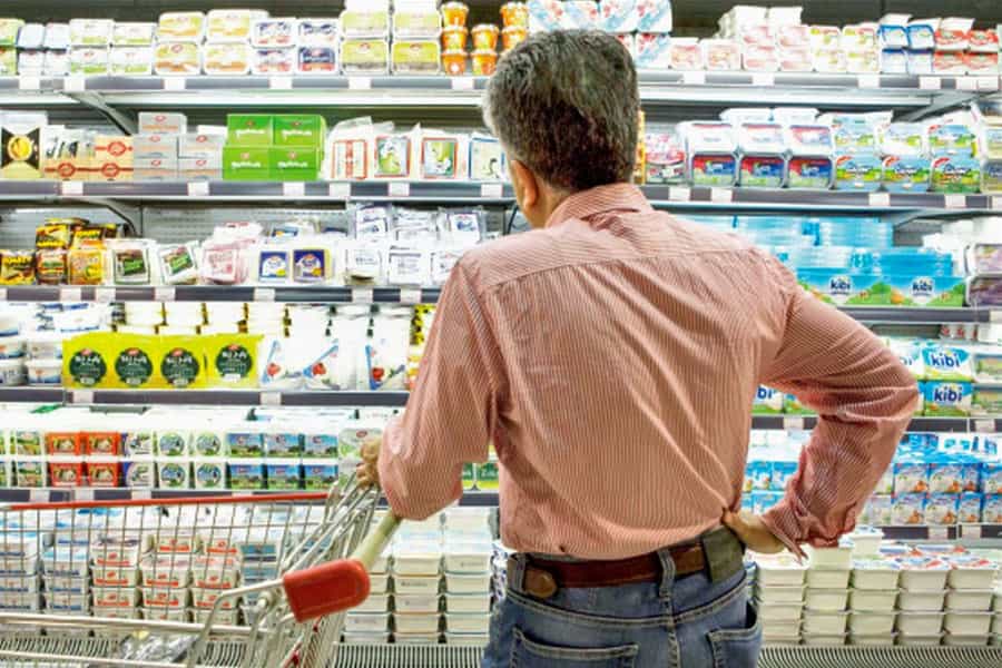 مردی با چرخ دستی خرید روبه‌روی یخچال اجناس لبنی یک فروشگاه ایستاده و به اجناس نگاه می‌کند.