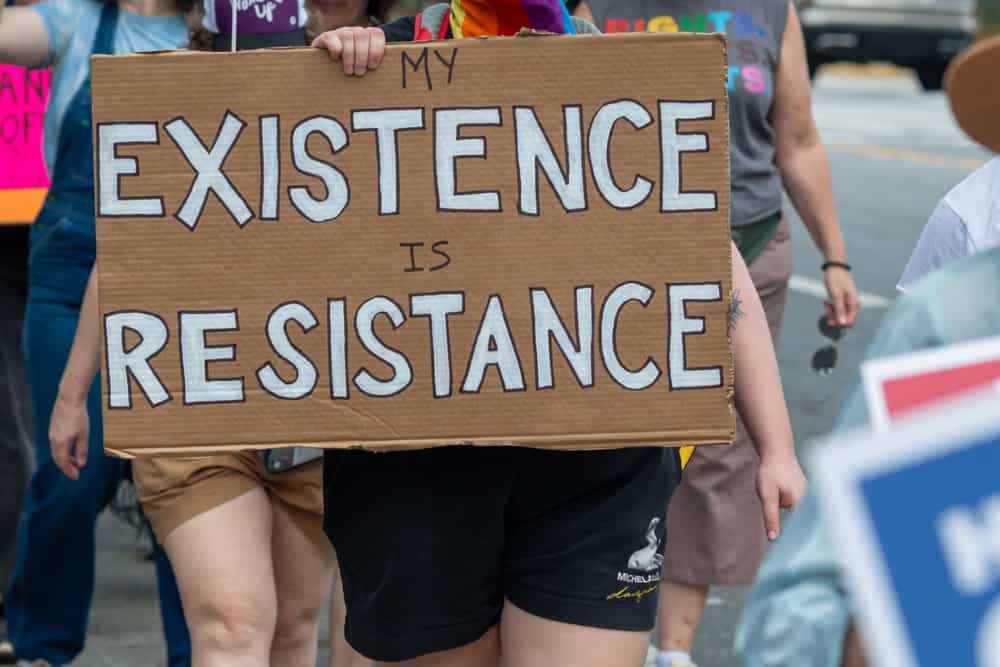 فردی در راهپیمایی «دستها دور» در ۵ آوریل ۲۰۲۵ در آتلانتا، جورجیا، تابلویی در دست دارد که روی آن نوشته شده «بودن من مقاومت است».