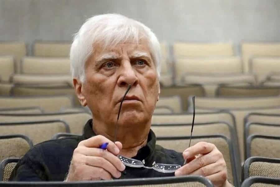 بهرام بیضایی در سالن تئاتر نشسته و خودکار و عینک خود را در دست دارد.