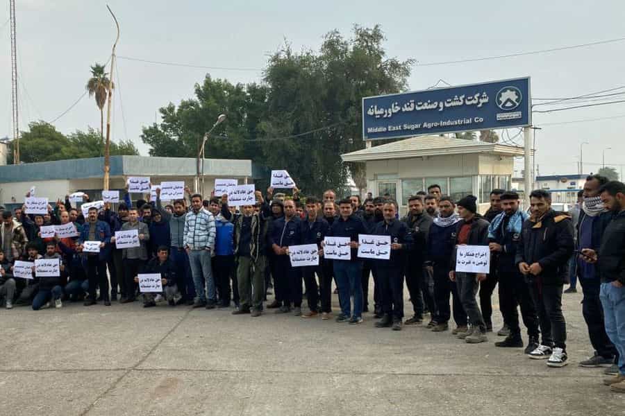 کارگران اعتصابی شرکت قند خاورمیانه شوش در مقابل ورودی کارخانه ایستاده‌اند و پلاکاردهایی که خواسته‌هایشان را بازتاب می‌دهند در دست دارند.