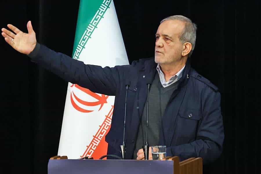 مسعود پزشکیان پشت تریبون یک دست خود را بلند کرده و در حال سخنرانی است. پرچم جمهوری اسلامی هم پشت سر او دیده میشود.