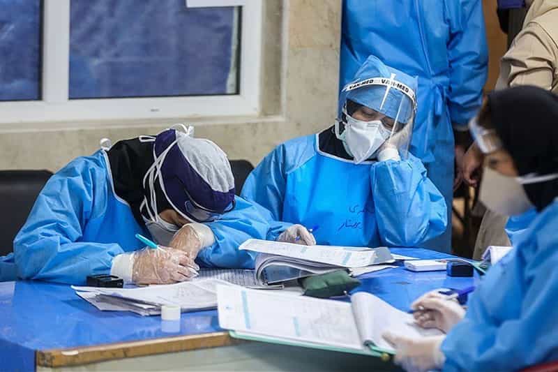 چند پرستار با لباس و ماسک پشت یک میز خسته نشسته‌اند.