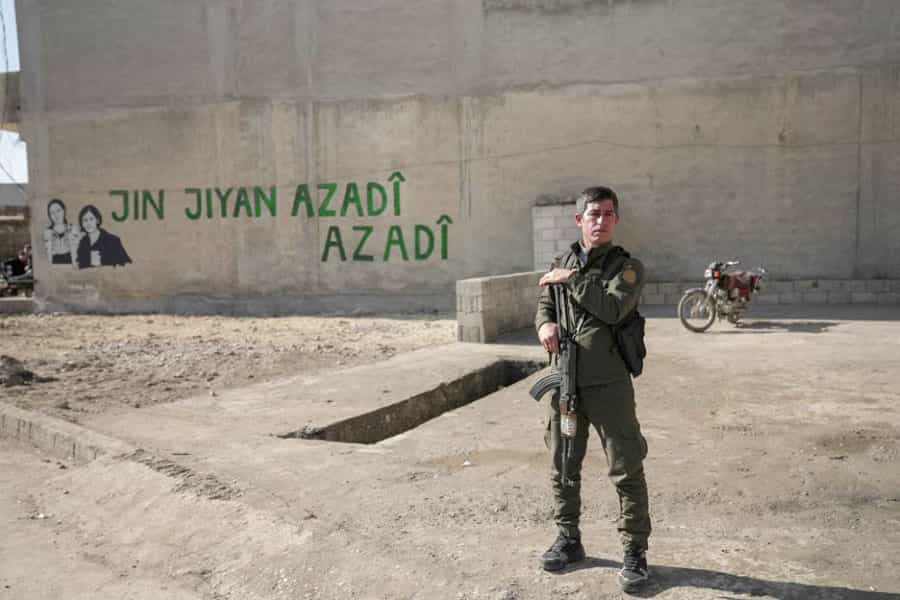 یکی از اعضای نیروهای دموکراتیک سوریه (SDF) در حسکه سوریه، در مقابل دیواری که روی آن نوشته شده: «ژن، ژیان، ئازادی» (زن، زندگی، آزادی) ایستاده است