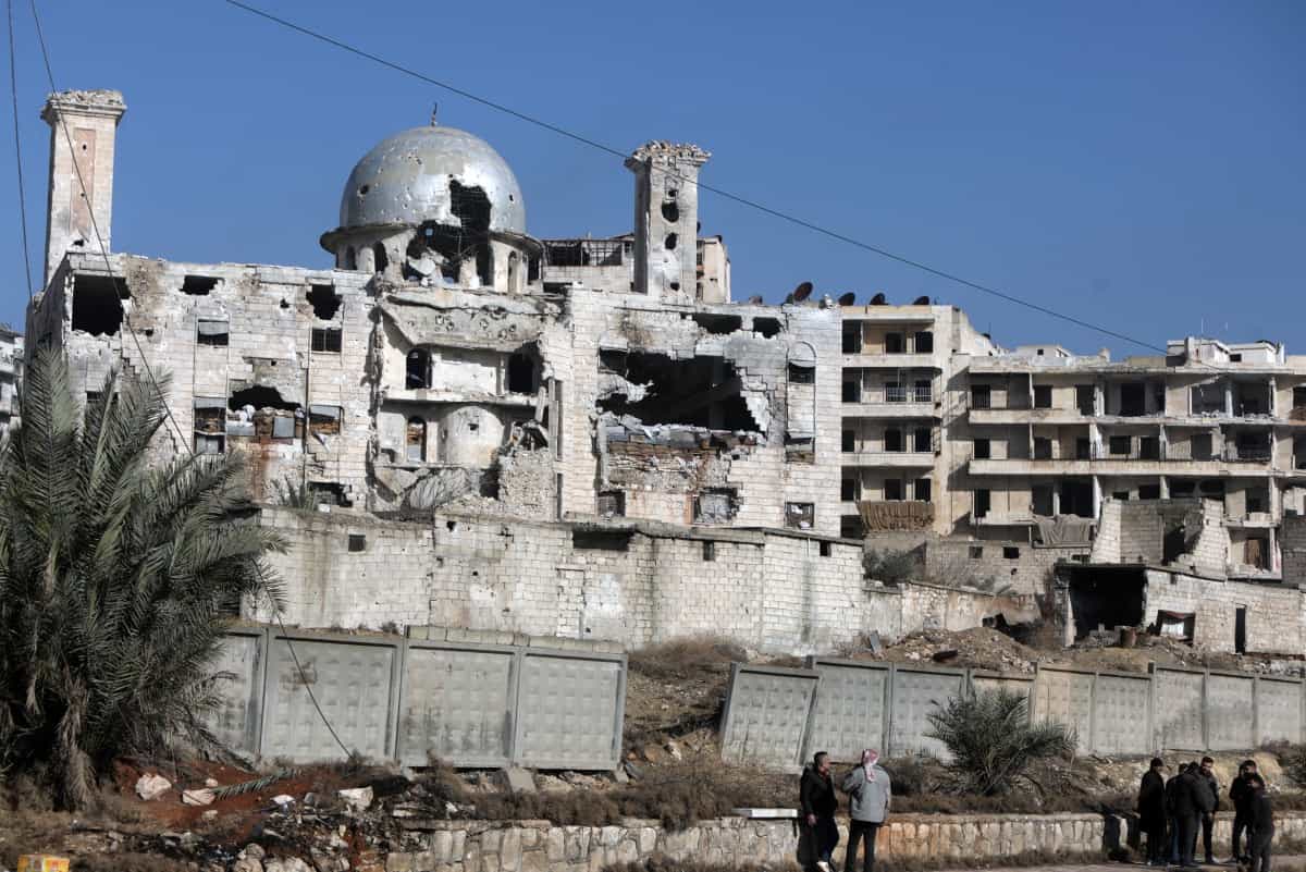 ساکنان محله شیخ مقصود در پسزمینه ساختمانهای آسیبدیده، در حلب، ۱۰ ژانویه ۲۰۲۶، پس از درگیریهای سنگین بین نیروهای امنیتی دولت سوریه و نیروهای کرد ایستادهاند.