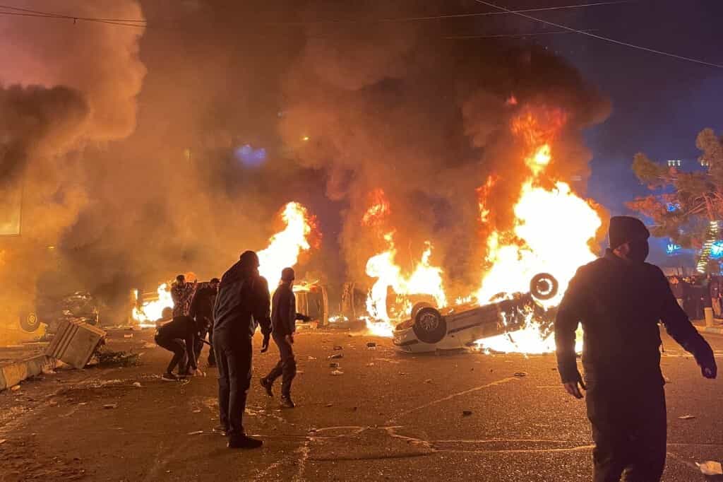 تظاهرکنندگان ایرانی در اطراف خودروهای در حال سوختن در نزدیکی یک مسجد تجمع کرده و خیابانی را در جریان اعتراضات در تهران، ایران، در 8 ژانویه 2026 مسدود کردند.