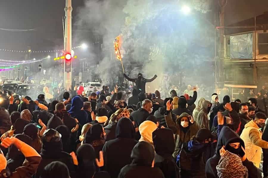 در هوای تاریک شب یکی از معترضان که چهره خود را پوشانده است چوبی را آتش زده و آن را در مقابل جمعیتی که روبه‌رویش ایستاده، بلند کرده است. دست دیگرش را مشت کرده و بالا آورده است.