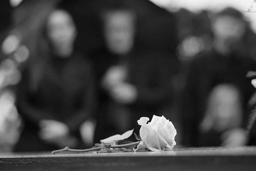 گل سفیدی بر مزار یک نفر و افرادی سیاه پوش در پسزمینه تصویر.