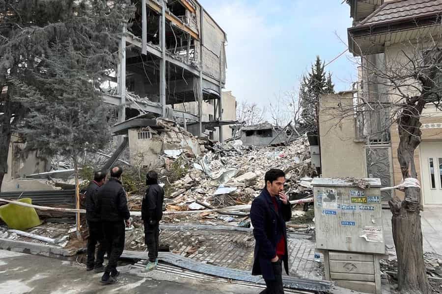 در یک کوچه خانه‌ای ویران شده و چند نفر در کنار آن ایستاده‌اند یا از برابر آن عبور می‌کنند.