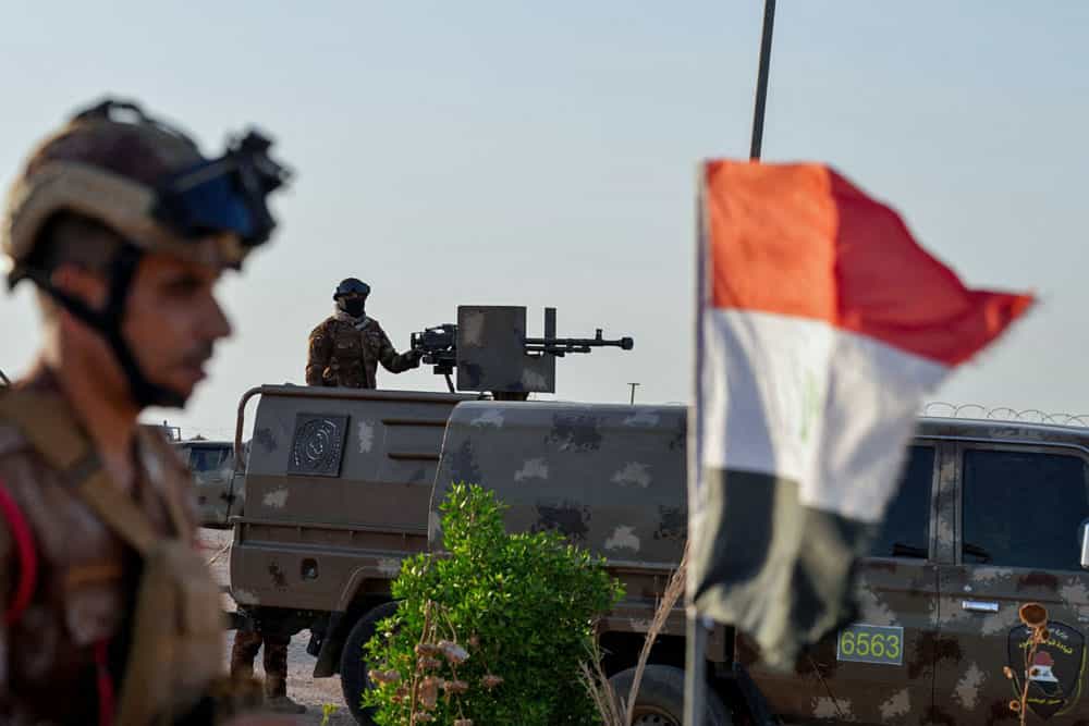در تصویر دو سرباز عراقی در کنار پرچم این کشور دیده می‌شوند. یکی از آن‌ها پشت یک تیربار بر روی یک خودرو نظامی است.