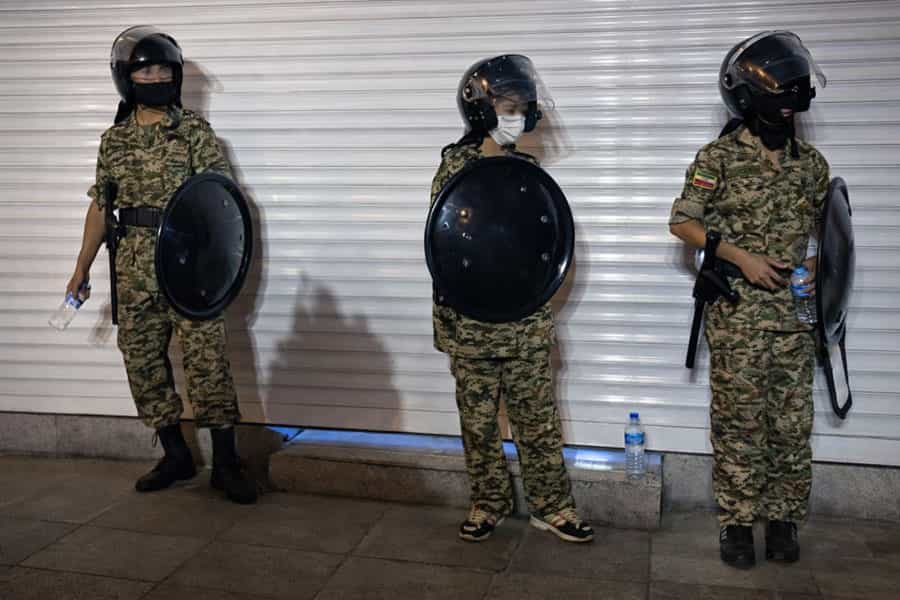 سه کودک بسیجی با سپر و کلاهخود ضدشورش و لباس نظامی در کنار کرکره یک مغازه ایستادهاند.