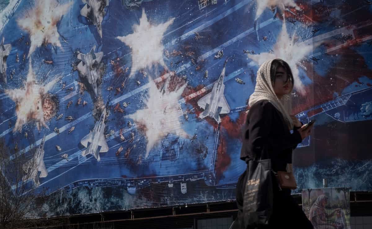 زن جوانی ایرانی از مقابل ساختمانی دولتی در مرکز تهران عبور می‌کند که با یک بیلبورد عظیم ضدآمریکایی پوشیده شده و تصویر نمادین ناو هواپیمابر USS Abraham Lincoln (CVN-72) را در حال نابودی نشان می‌دهد. این صحنه در روز ۲۶ فوریه ۲۰۲۶، آخرین روز مذاکرات ایران و آمریکا در شهر ژنو ثبت شده است. (عکس: مرتضی نیکوبذل / نورفوتو) (عکس: مرتضی نیکوبذل / نورفوتو via AFP)