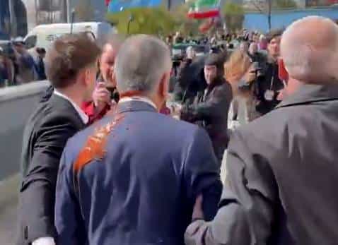 تصویر رضا پهلوی را از پشت سر نشان می‌دهد که مایع قرمزرنگ به روی او پاشیده شده است. دو مأمور امنیتی در کنار او قرار دارند.