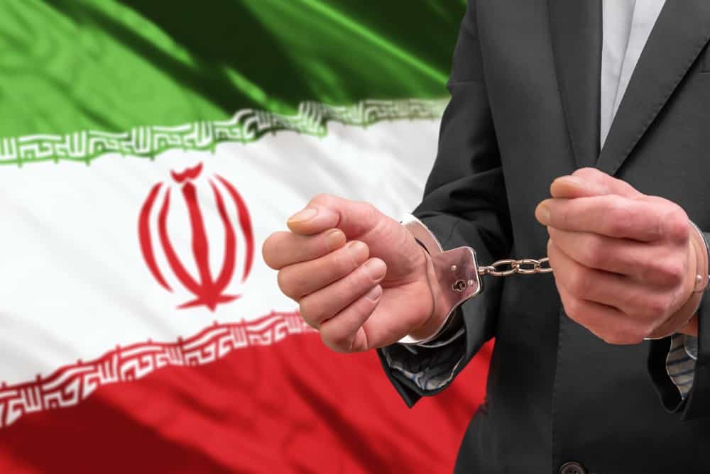 تصویری از دو دست که دستبند خوردهاند و پرچم جمهوری اسلامی ایران در پسزمینهی آن