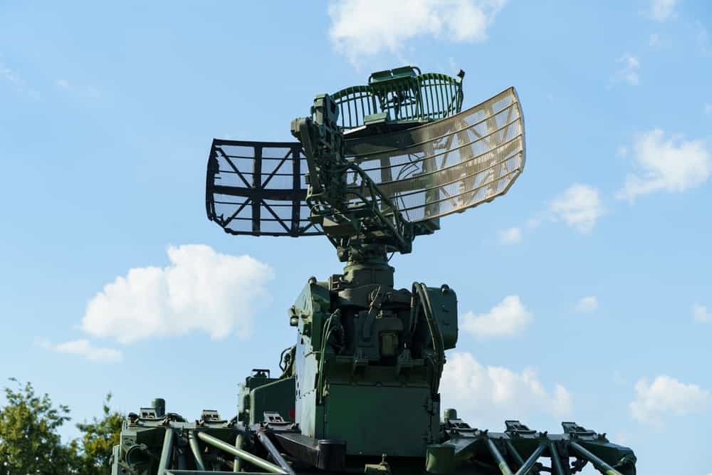 یک سیستم رادار نظامی در مقابل پس‌زمینه‌ای از ابرهای پف‌دار ایستاده و قطعات مکانیکی پیچیده‌ای را به نمایش می‌گذارد. این فناوری پیشرفته به نظارت و شناسایی در طول عملیات کمک می‌کند.