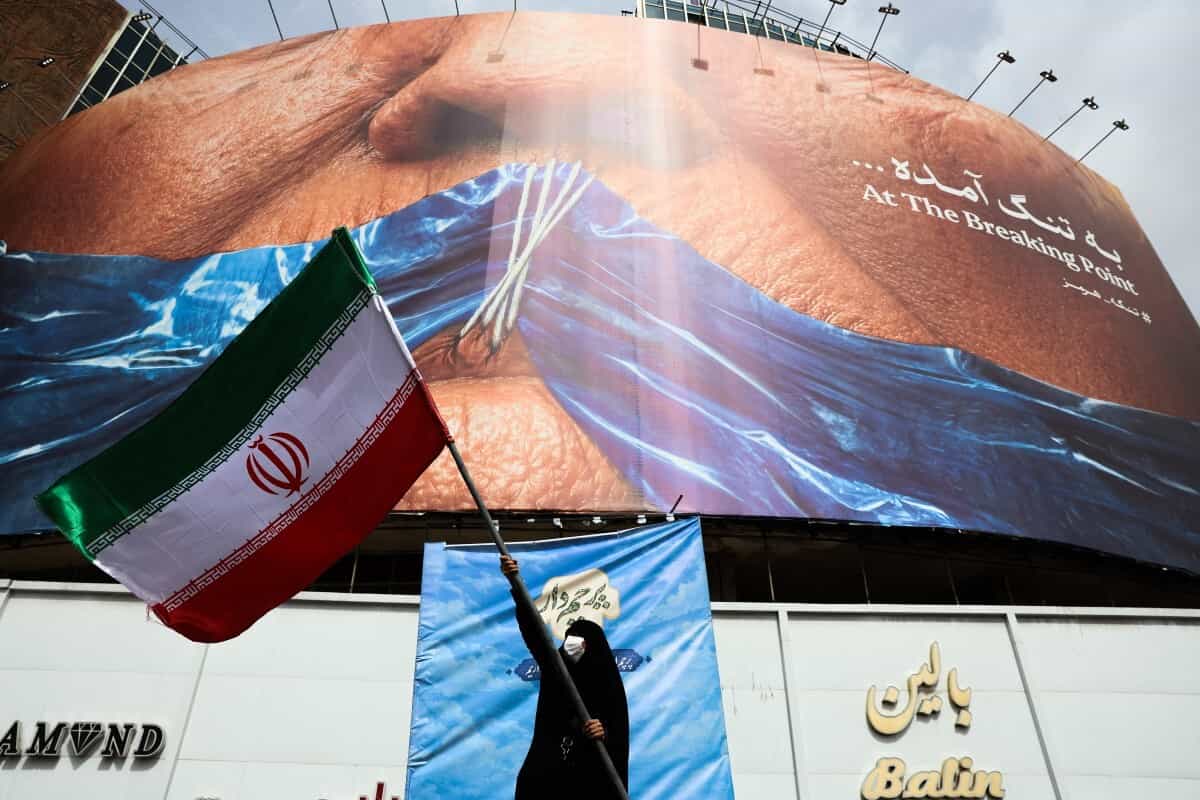 زنی در میدان ولیعصر تهران، ۵ مه ۲۰۲۶، پرچم ایران را در مقابل یک بیلبورد ضد آمریکایی که به دونالد ترامپ، رئیس جمهور آمریکا و تنگه هرمز اشاره دارد، تکان می‌دهد.