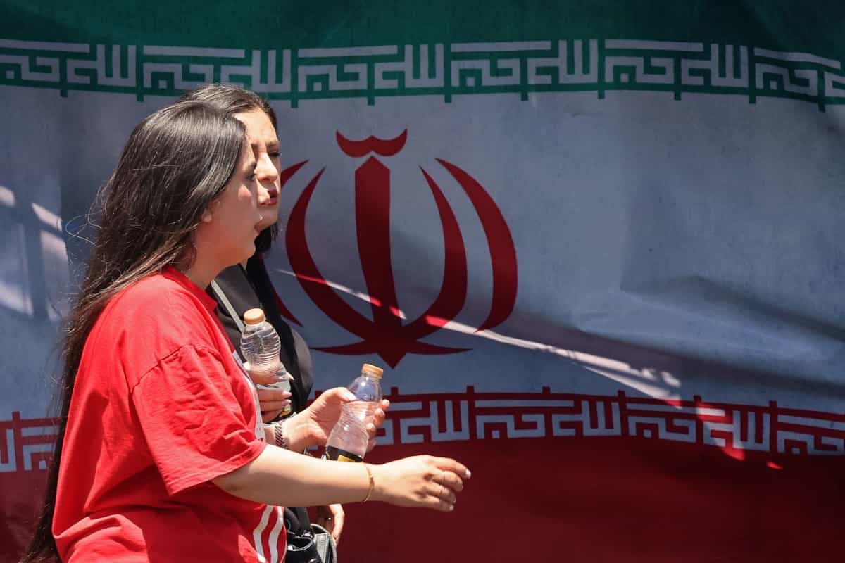 دو زن بیحجاب در حال گفتوگو با هم در خیابان از کنار پرچم جمهوری اسلامی ایران عبور میکنند.