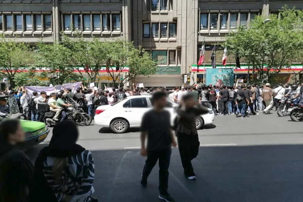 در یک هوای آفتابی دانشآموزان معترض در مقابل وزارت آموزش و پرورش تجمع کردهاند و خودروها از خیابان عبور میکنند.