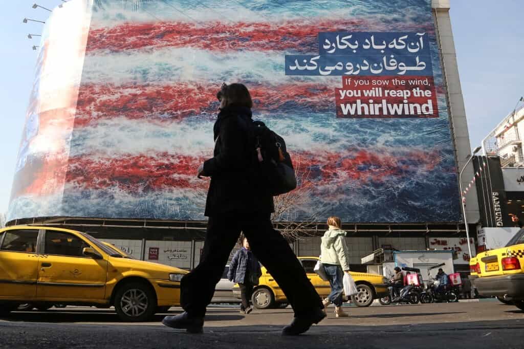 تهران، ایران - ۲۸ ژانویه: مردم در پی مداخله احتمالی ایالات متحده علیه ایران در ۲۸ ژانویه ۲۰۲۶ در تهران، ایران، از کنار یک پوستر دیواری ضد آمریکایی که روی آن نوشته شده «اگر باد بکارید، گردباد درو خواهید کرد» عبور میکنند.