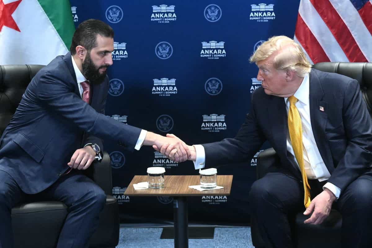 ترامپ و الشرع دست یکدیگر را فشردهاند و پشت سر آنها پرچمهای آمریکا و سوریه دیده میشود.