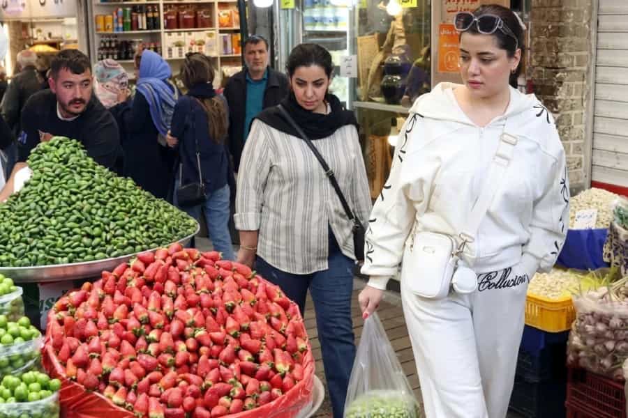 دو زن در بازار میوه و سبزیجات دیده میشوند. فروشنده و عدهای دیگر پشت سر آنها هستند