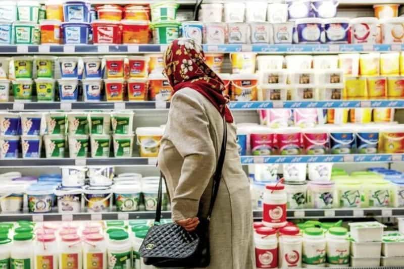 یک زن سالمند روبه‌روی یخچال یک فروشگاه ایستاده و به اجناس نگاه می‌کند.
