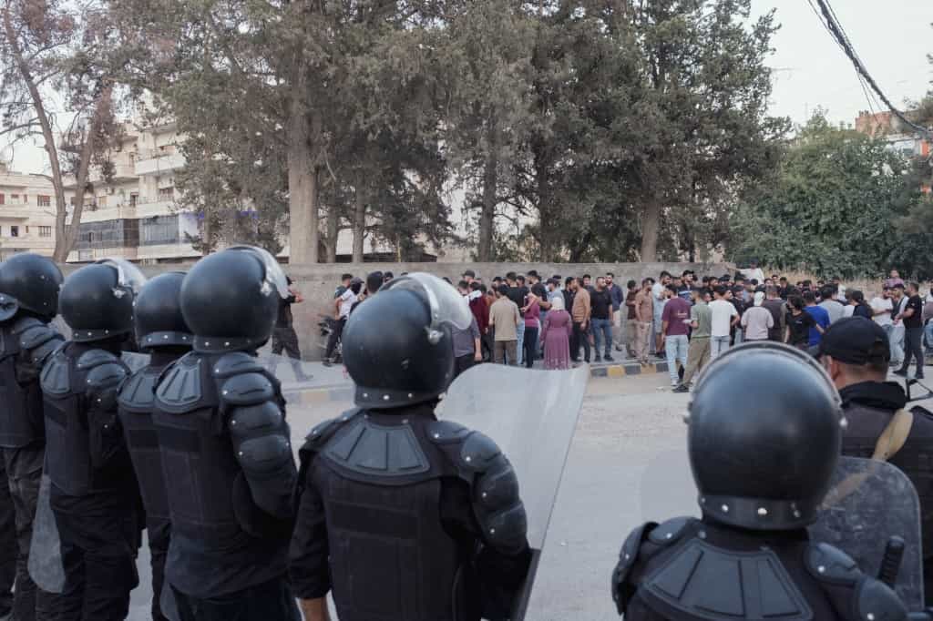 در این تصویر در یک طرف خیابان معترضان قرار دارند و در سوی دیگر مأموران امنیتی سوریه با کلاهخود بر سر.