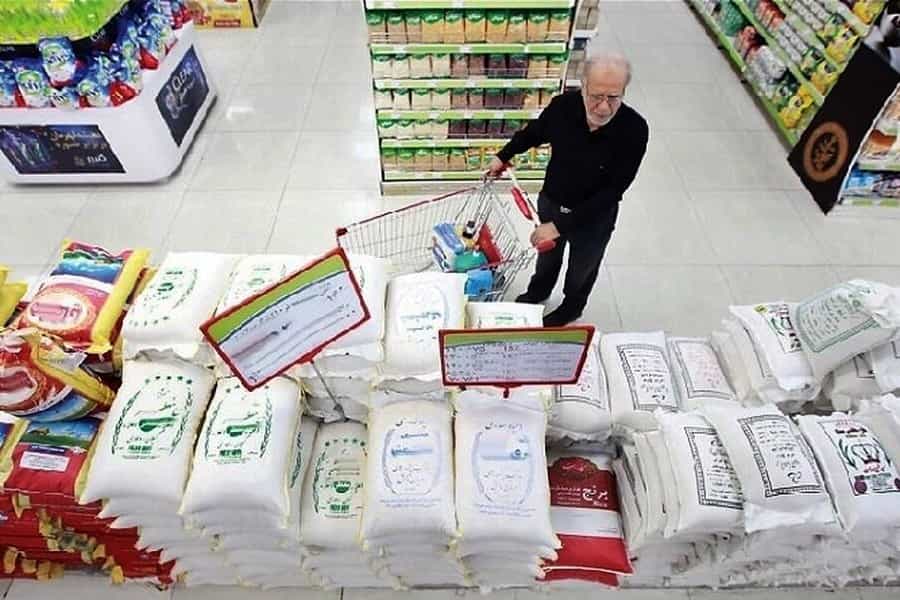مرد سالمندی در یک فروشگاه در مقابل کیسههای برنج با مارکهای مختلف ایستاده و به قیمتهای آنها نگاه میکند.